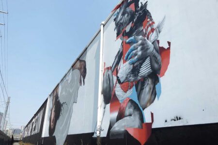 A long mural on a building wall features dynamic, fragmented images of human figures and abstract elements, painted in vibrant colors against a pale background. The artwork stretches along a pathway under a clear blue sky.