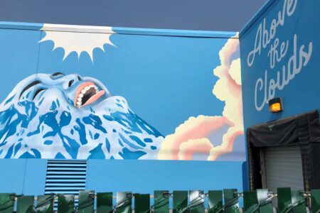 A vibrant mural on a blue building depicts a stylized face with open mouth, blending into snowcapped mountains and clouds. Sunlight shines above. The words "Above the Clouds" are painted on the upper right. Green fence is in the foreground.
