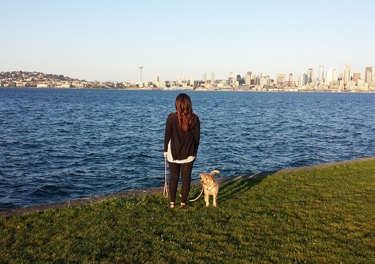 A person with long hair stands on a grassy shoreline, facing a large body of water with a city skyline in the background. A dog on a leash stands beside them, looking in the same direction.