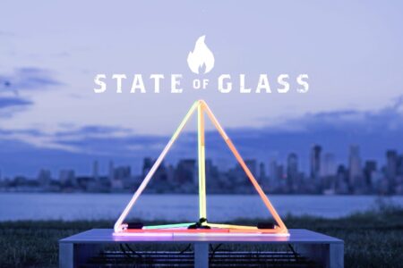 A glowing neon triangle sculpture sits on a platform with a city skyline and water in the background. Above the sculpture, the text reads "State of Glass." The sky is a dusky blue-gray, suggesting evening or twilight.