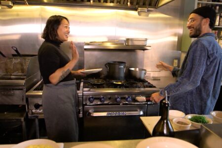 Two people are smiling and talking in a professional kitchen. One person holds a plate, and the other gestures with their hands. Pots and pans are on the stovetop, and various bowls and ingredients are on the counter in the foreground.