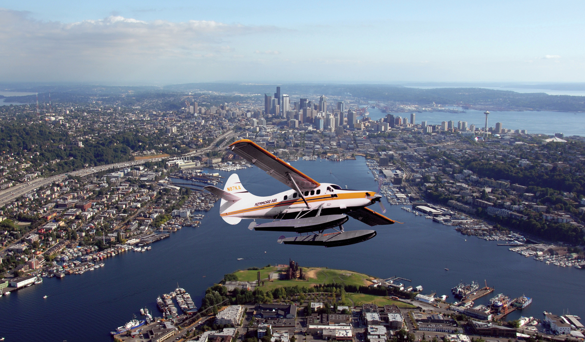A small floatplane flies over a cityscape with waterways and green areas below, while a downtown skyline with tall buildings is visible in the background under a partly cloudy sky.