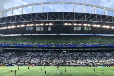 A wide-angle view of a soccer stadium during a match, with players on a green field. The stands are filled with spectators, and a large arching roof covers the stadium. The sky is partly cloudy.