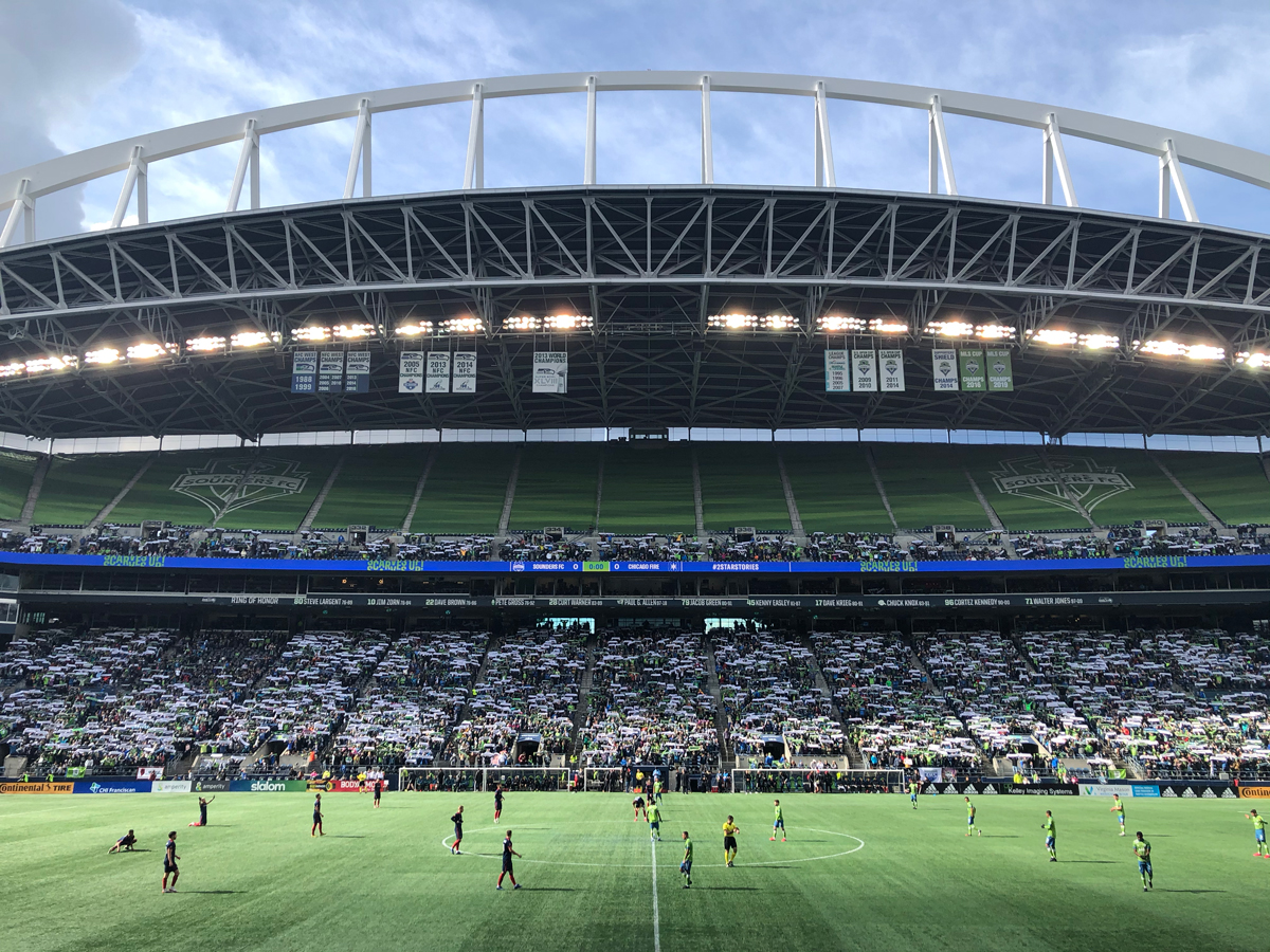 A wide-angle view of a soccer stadium during a match, with players on a green field. The stands are filled with spectators, and a large arching roof covers the stadium. The sky is partly cloudy.