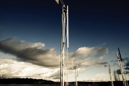 Tall, modern wind turbines stand in an open field under a dramatic sky with dark clouds and a hint of sunlight. A body of water and distant hills are visible in the background, creating a serene and dynamic landscape.