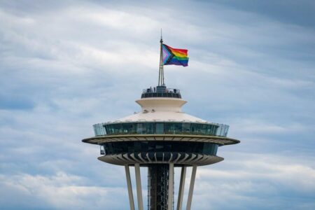 Seattle's Space Needle with a rainbow flag flying at the top against a backdrop of a cloudy sky.