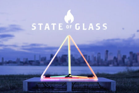 A neon-lit geometric sculpture in the shape of a triangle sits on a platform by a waterfront. The words 