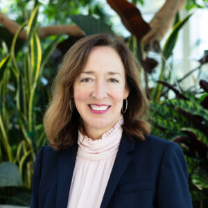 A photo of a woman with short brown hair wearing a pink high-neck blouse under a black blazer. She stands in front of green plants and a tree with tree brown trunk.