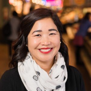 A person with shoulder-length dark hair and a bright smile is wearing a white scarf with black polka dots and a black top. The background is softly blurred, suggesting an indoor setting with warm lighting.