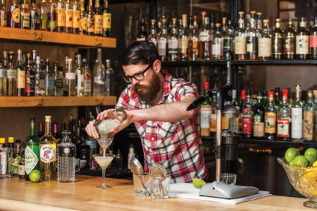 A bearded bartender in a plaid shirt pours a cocktail into a glass from a shaker. The bar behind him is stocked with various bottles of liquor and mixers. A lime squeezer and fresh limes are visible on the wooden counter.
