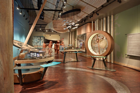 A modern museum exhibit with wooden accents displays artifacts, informational panels, and circular installations. The room features high ceilings and various indigenous art pieces, highlighted by soft lighting.