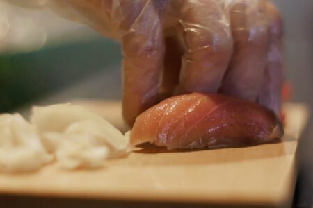 A gloved hand carefully handles a piece of raw tuna on a wooden cutting board, with slices of fresh seafood nearby. The focus is on the fish and the hand, suggesting food preparation or sushi making.