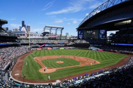An aerial view of a baseball game at a stadium with a partially open roof. The field is bright green, with players positioned around the bases. The stands are filled with spectators, and a city skyline is visible in the background under a clear blue sky.