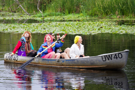 Three people in colorful clothing are enjoying a canoe ride on a serene lake surrounded by greenery and lily pads. They appear happy, with one person paddling while the others relax and smile.