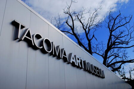 The image shows the exterior of the Tacoma Art Museum with large, metallic letters on a sleek facade. Leafless trees stand against a backdrop of blue sky with clouds, creating a stark contrast with the museum's modern design.