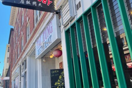 Street view of a red-brick building featuring a vertical green slat facade. Signs read "Tai Tung Chop Suey" and "Tai Tung Restaurant." A door with "OPEN" in neon is visible, alongside Chinese characters.
