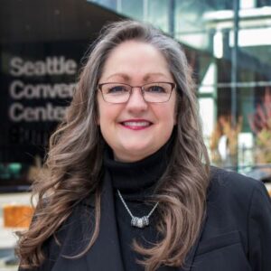 A smiling woman with long gray hair and glasses stands in front of a building. She is wearing a black turtleneck and a necklace, with a black blazer. The words 