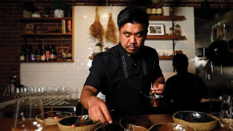 A chef in a black uniform carefully plating food in a dimly lit restaurant kitchen, with shelves of bottles, glasses, and hanging herbs visible in the background.