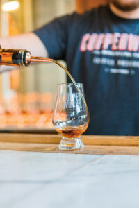 A person in a dark shirt is pouring liquor from a bottle into a glass labeled "Copperworks." The glass is on a wooden surface, and the background is softly blurred.
