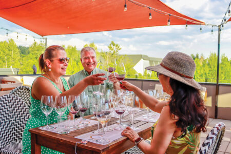 A group of three people sit at a wood table on a patio under an orange umbrella. They cheers their wine glasses. Several wine glasses fill the table for their wine tasting.