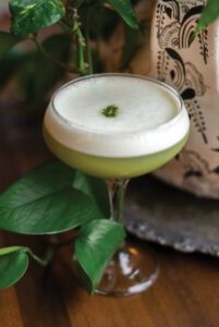 A frothy, light green cocktail in a stemmed glass sits on a wooden table, garnished with a small green herb. Lush green leaves surround the glass, while a decorative black and white patterned vase is partially visible in the background.