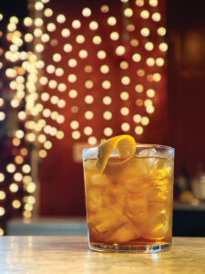 A glass of iced tea with an orange twist sits on a table. The background features blurred, circular yellow lights against a red wall, creating a warm and inviting atmosphere.