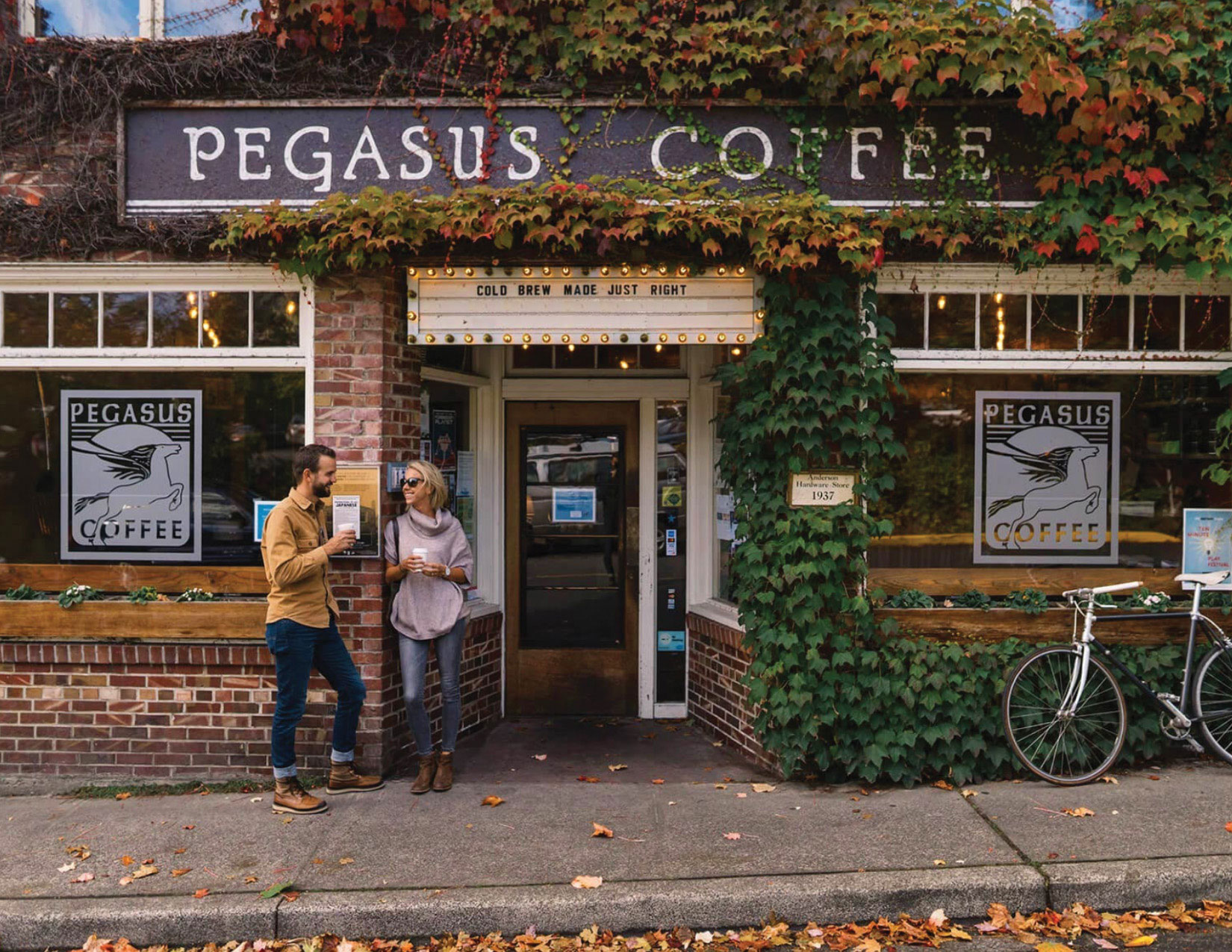 A man and woman stand outside Pegasus Coffee, a brick building adorned with lush green ivy. They hold drinks and engage in conversation. A bicycle is parked nearby, and fallen leaves scatter the sidewalk. A sign reads, "Cold Brew Made Just Right".