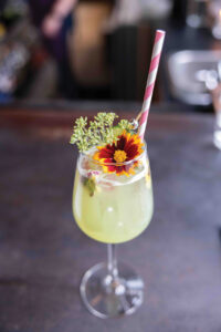 A refreshing green cocktail served in a wine glass, garnished with a vibrant orange and yellow flower and delicate greenery. A red and white striped straw completes the presentation. The drink is set on a dark table.
