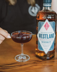 A close-up of a glass filled with a dark cocktail garnished with a cherry on a toothpick. A person with painted nails holds the glass. A bottle of Westland American Single Malt Whiskey is placed next to the glass on a wooden table.