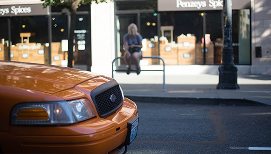 An orange taxi is in the foreground on a city street, with a store named "Penzeys Spices" in the background. A person is sitting on a railing near the shop. The scene is in daylight.