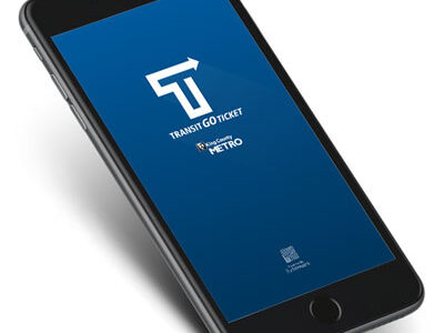 A smartphone displaying the Transit Go Ticket app on its screen, featuring the logo and text “Transit Go Ticket” with “Metro” underneath. The phone is tilted slightly and set against a white background.