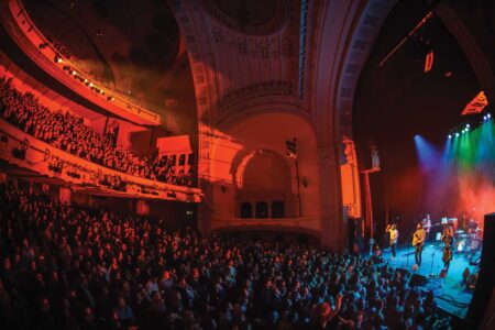 A wide-angle view of a concert in a grand theater, showcasing a band performing on stage with colorful lighting. The audience fills the seats and stands, enjoying the lively atmosphere under the ornate architectural details of the venue.