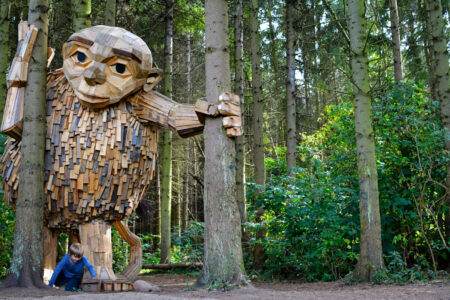A large wooden troll sculpture stands in a forest, its hand resting on a tree. A child plays at its feet, surrounded by tall trees and green foliage.