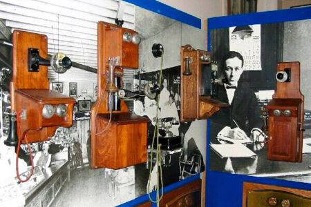 A display of vintage wooden wall-mounted telephones against a backdrop featuring a black and white historical photograph of a man writing at a desk.