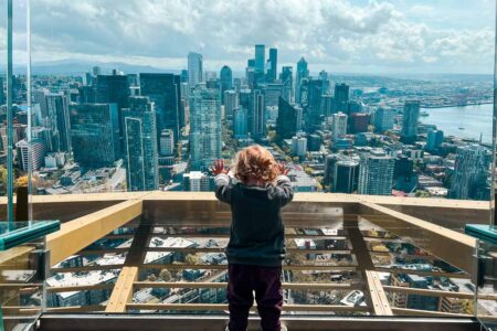 A small child standing in front of large glass window at the top of the Space Needle overlooking the cityscape.