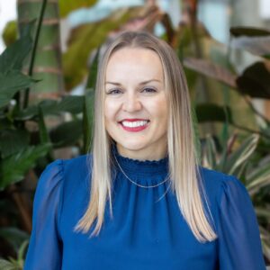 A person with long blonde hair, wearing a blue blouse and gold necklace, smiling while standing in front of lush green plants.