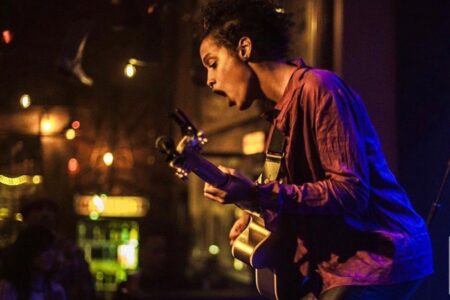 A musician passionately performs on stage, playing an acoustic guitar. Warm lights create a cozy atmosphere. The performer wears a red shirt and leans forward, deeply immersed in the music.