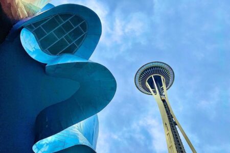 View of the Seattle Space Needle from below under a cloudy sky, with the curving, reflective metal facade of the Museum of Pop Culture in the foreground. The structures create a dynamic contrast against the light blue background.