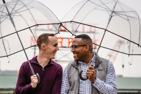 Two people stand outdoors under clear umbrellas, smiling at each other. The background features a blurred sign and overcast sky. They wear casual, warm clothing, conveying a sense of joy and togetherness.