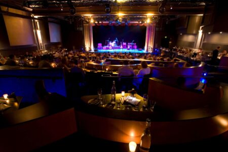 A dimly lit music venue with a stage ready for a performance. Tables are set with drinks and candles, and attendees are seated, facing the stage. The space has a warm ambiance with blue and yellow lighting highlighting the stage area.