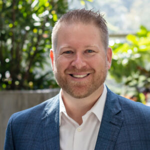 A smiling man with short hair and a beard, wearing a blue checked blazer and a white shirt. The background features lush green plants and soft natural lighting.