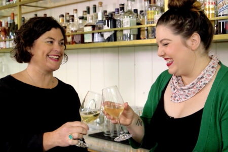 Chef Renee Erickson, wearing a black shirt, raises a glass with musician Mary Lambert, wearing a green cardigan over a black top and a white and red dotted scarf tied around her neck, as they stand in front of a bar lined with bottles of spirits.