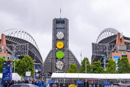 Lumen Field stadium with its distinctive arching roof structures, large central scoreboard, and crowds of people gathered outside on a cloudy day.