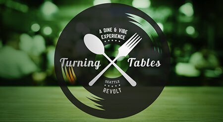 A logo with a black circular background featuring a spoon and fork crossed over each other. The text reads "A Dine & Vibe Experience" above "Turning Tables" and "Seattle Revolt" below. The background is a blurred setting with green tones.