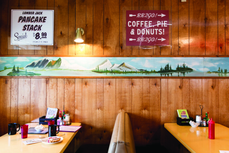 A cozy diner with wooden walls features a mural of mountains and trees. Booth seats have condiments and menus on the tables. Signs advertise pancake specials and coffee, pie, and donuts. Warm lighting creates a welcoming atmosphere.