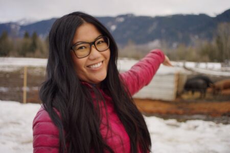 A woman with long black hair and glasses smiles at the camera while pointing towards the right. She is wearing a pink jacket. In the background, there is a snowy landscape with mountains and a few animals.