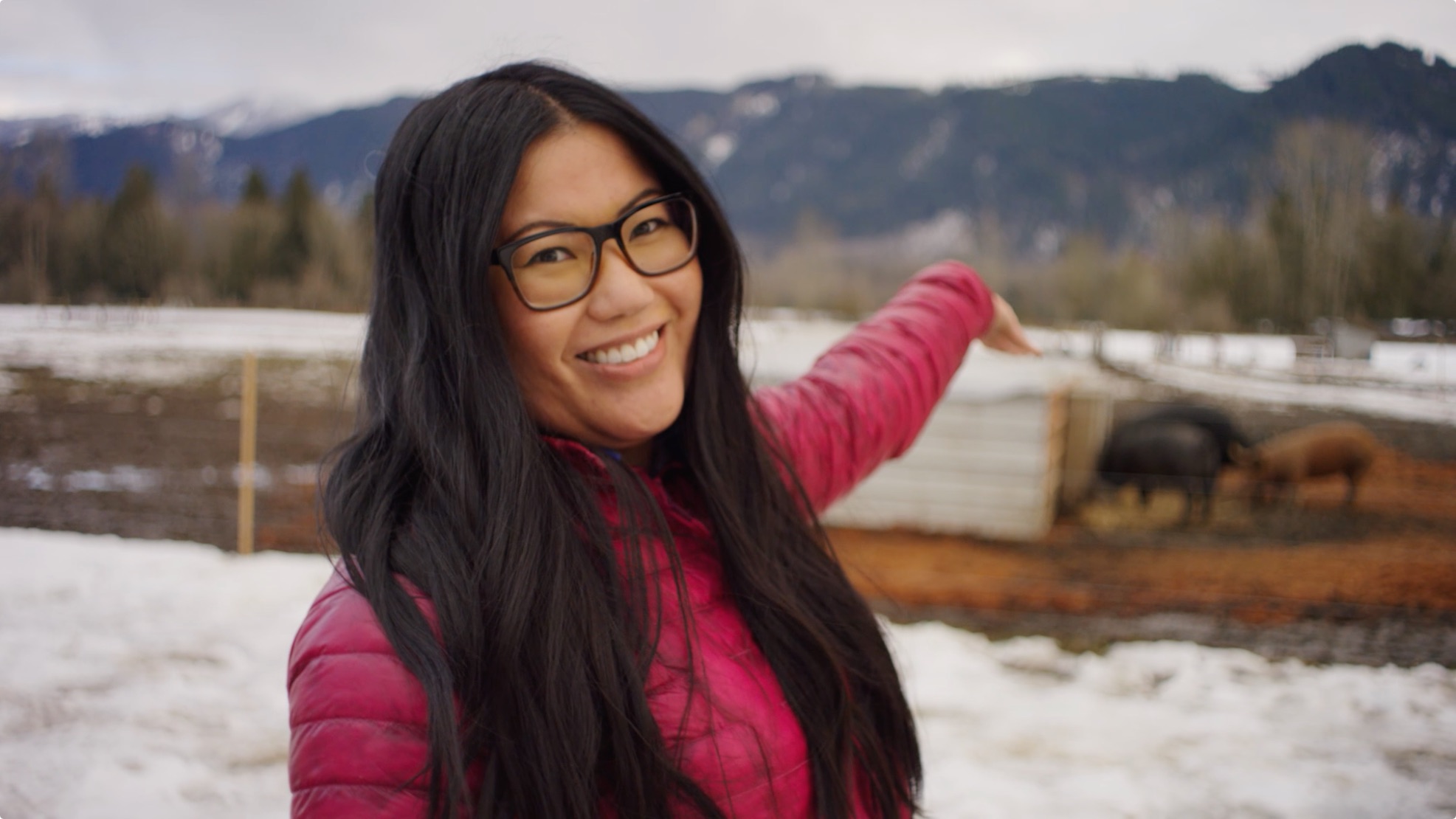 A woman with long black hair and glasses smiles at the camera while pointing towards the right. She is wearing a pink jacket. In the background, there is a snowy landscape with mountains and a few animals.