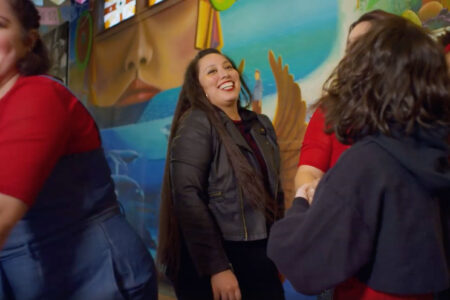 A group of people gather indoors in front of a colorful mural. One person in the center, smiling, wears a black jacket. Others, some in red, are engaged in conversation and laughter. The mural depicts a serene face and oceanic elements.