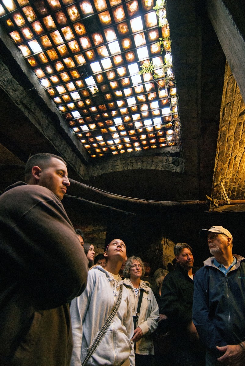 A group of people stand in a dim, underground room, looking up at a ceiling made of glass bricks that let light in from above. The walls are made of old, weathered bricks.