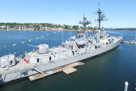 A large naval ship is docked at a harbor under clear blue skies. Its deck is adorned with colorful flags. In the background, there's a bridge and a landscape of trees and buildings. Nearby, a smaller boat is seen on the water.